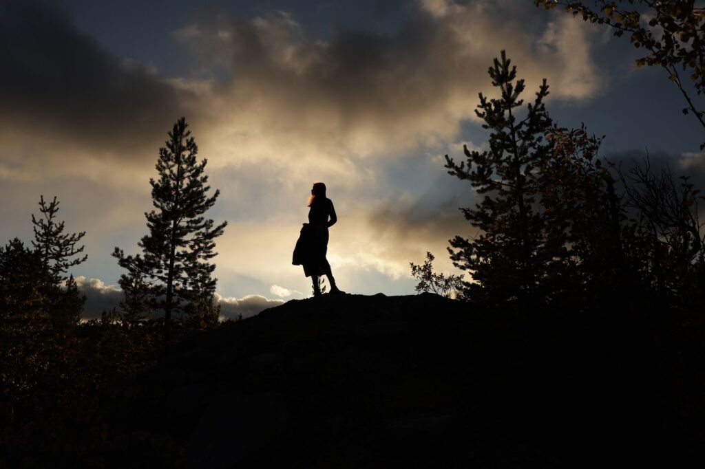 Nainen seisoo siluettina dramaattisen auringonlaskun taivasta vasten, puiden ympäröimänä.