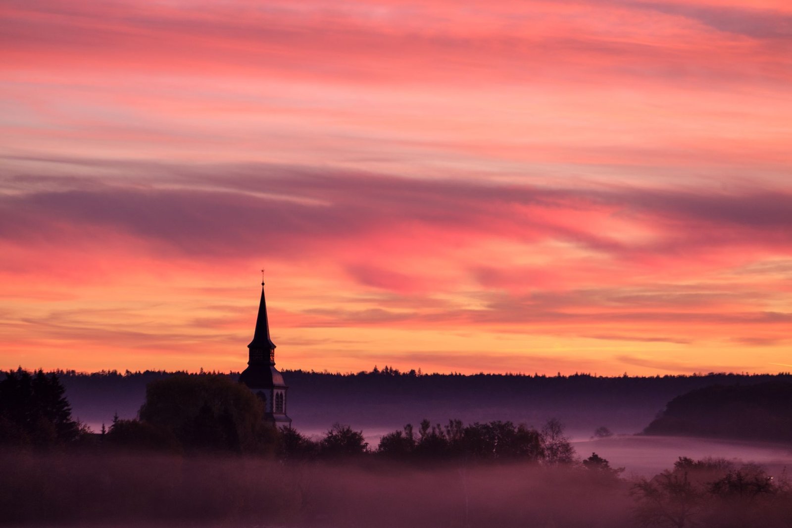 Maalaileva kirkontorni ilmestyy usvasta vankan auringonlaskun taivaan alla, luoden rauhallisen maiseman.