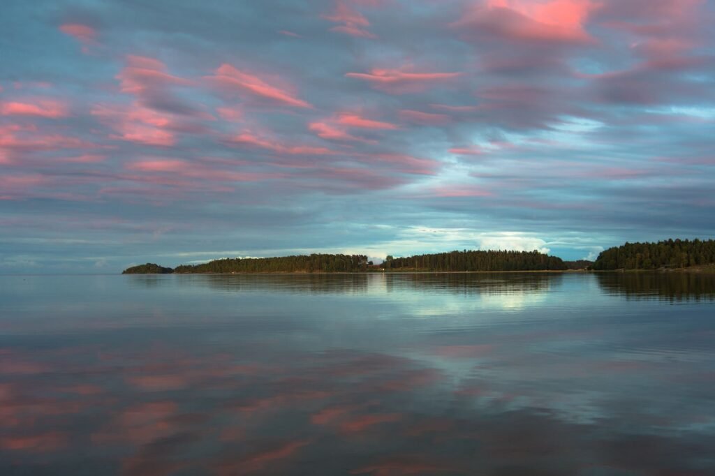 Rauhallinen auringonlasku järven yli, jossa vaaleanpunaiset pilvet heijastuvat vedessä, luoden rauhallisen maiseman.