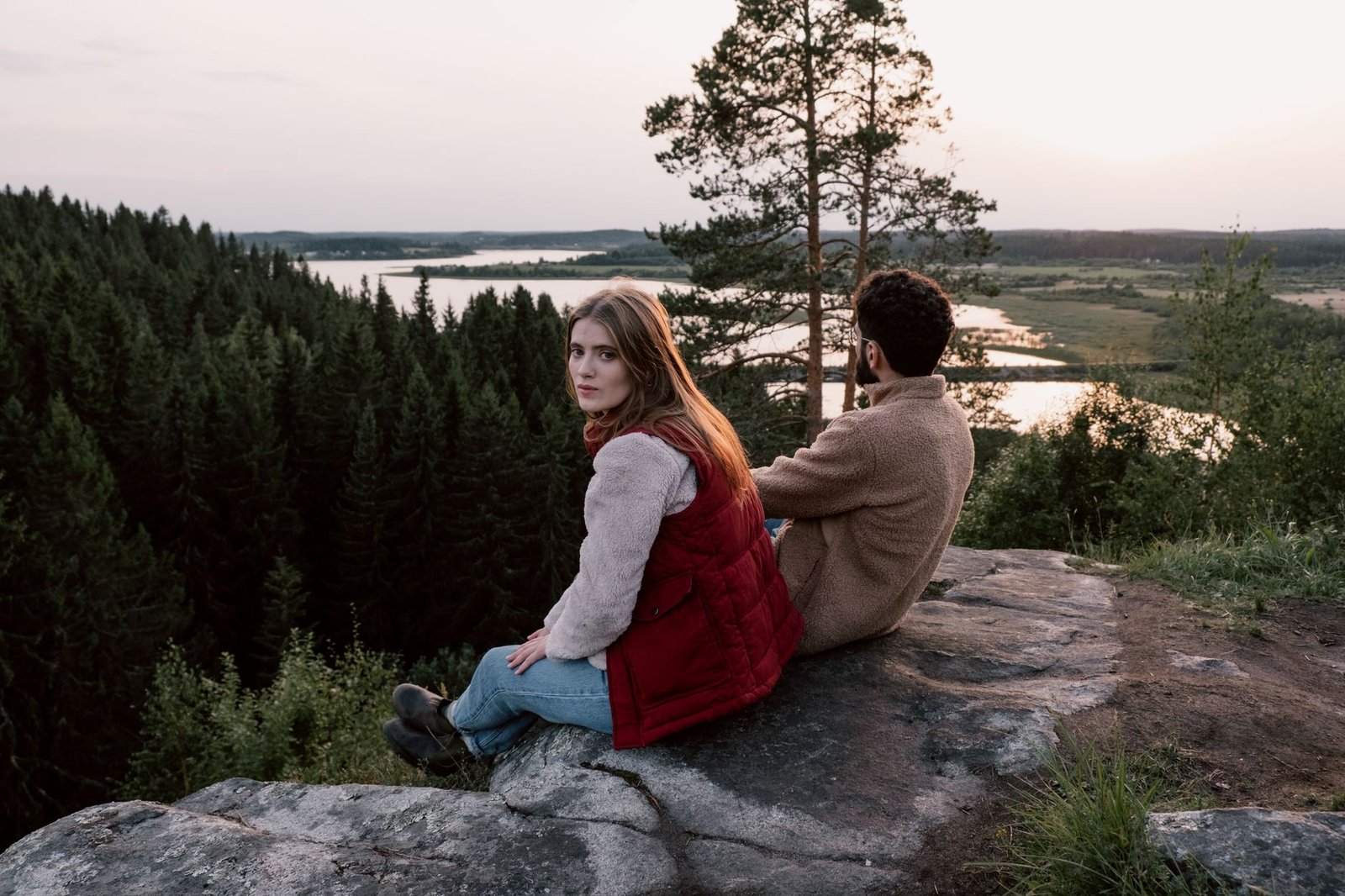 Pari istuu kivellä, katsellen rauhallista metsää ja jokea auringonlaskun aikaan.