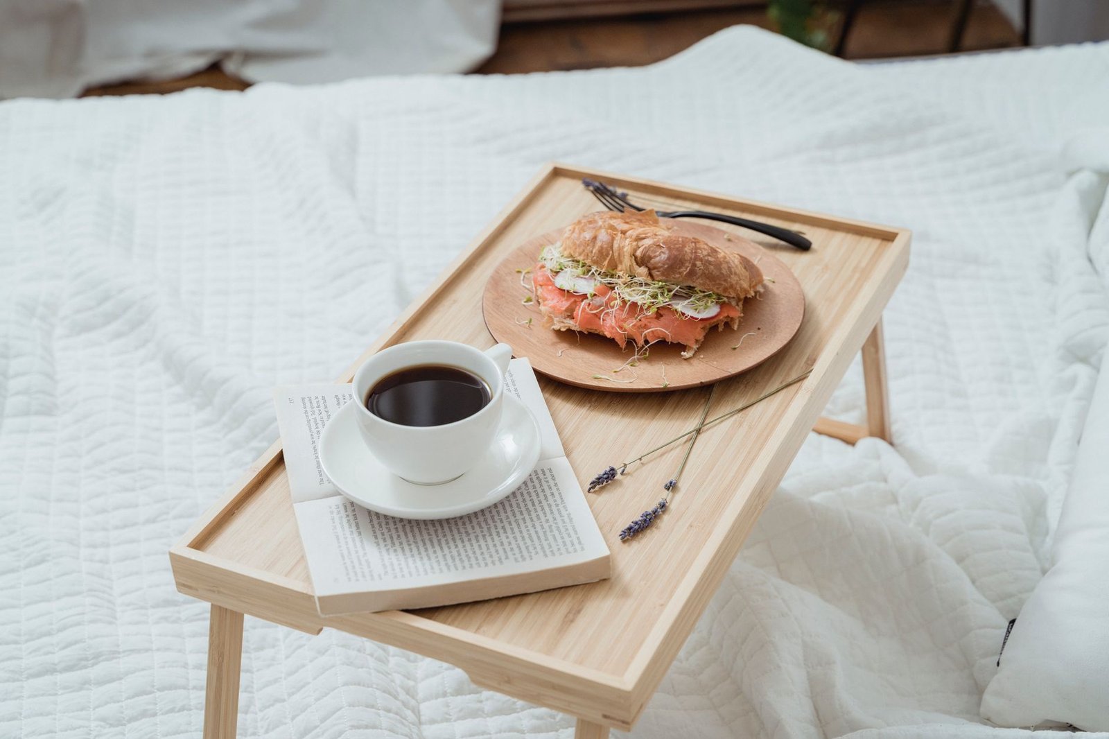 Ihana aamiainen sängyssä, jossa on kahvia ja croissant-sandwich puualustalla.
