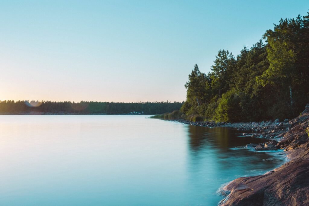Rauhallinen järvenranta, jossa on tyyni vesi ja runsas vihreä metsä kirkkaan taivaan alla.