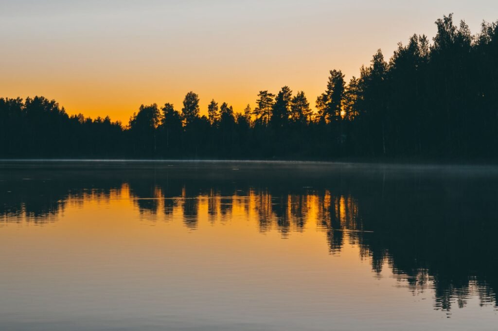 Rauhallinen aikainen aamunkoitto tyynellä järvellä Jyväskylässä, Suomessa, puiden siluetit heijastuvat vedessä.